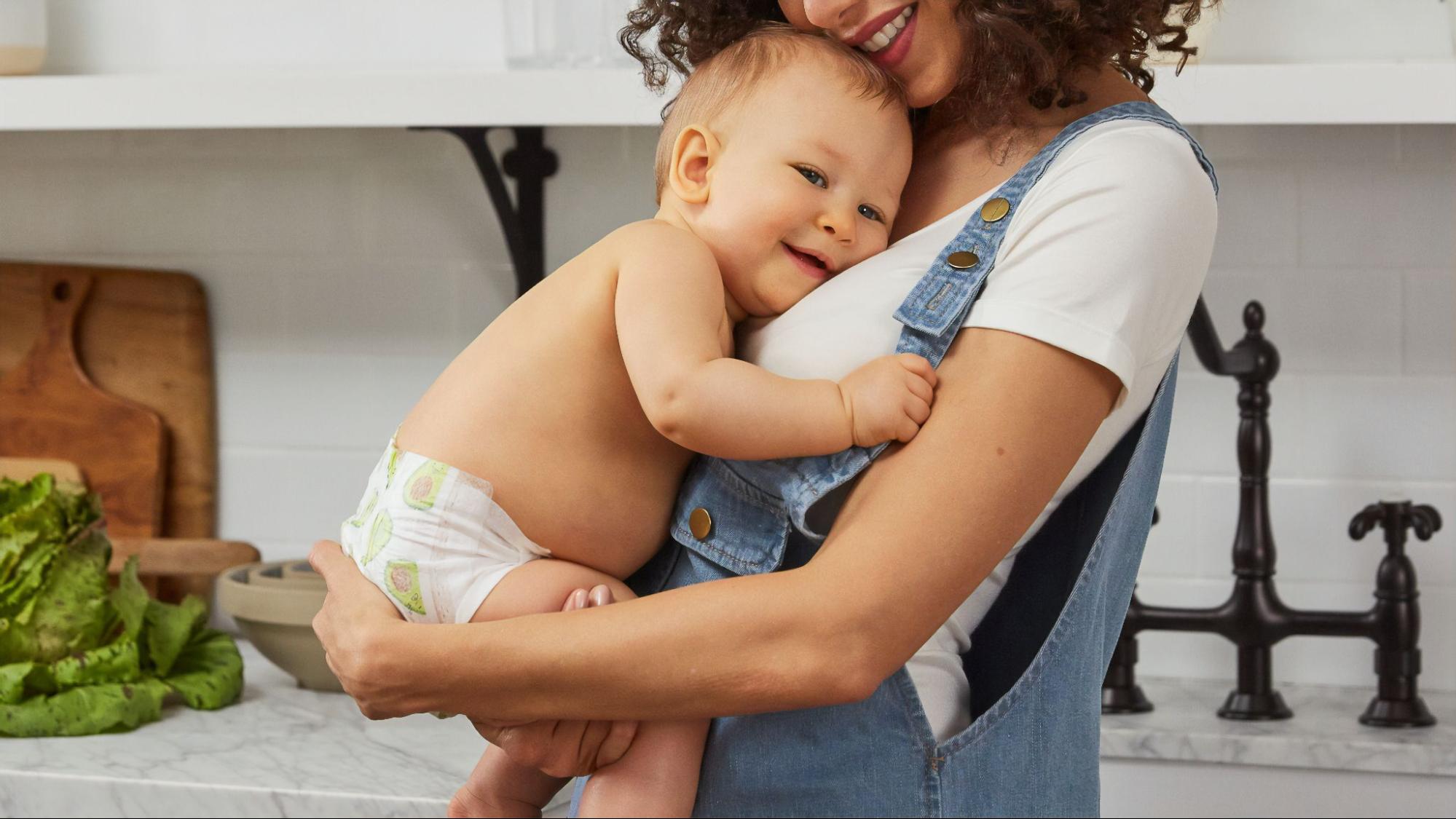 A smiling mother in overalls holding her baby in a shiny white kitchen.