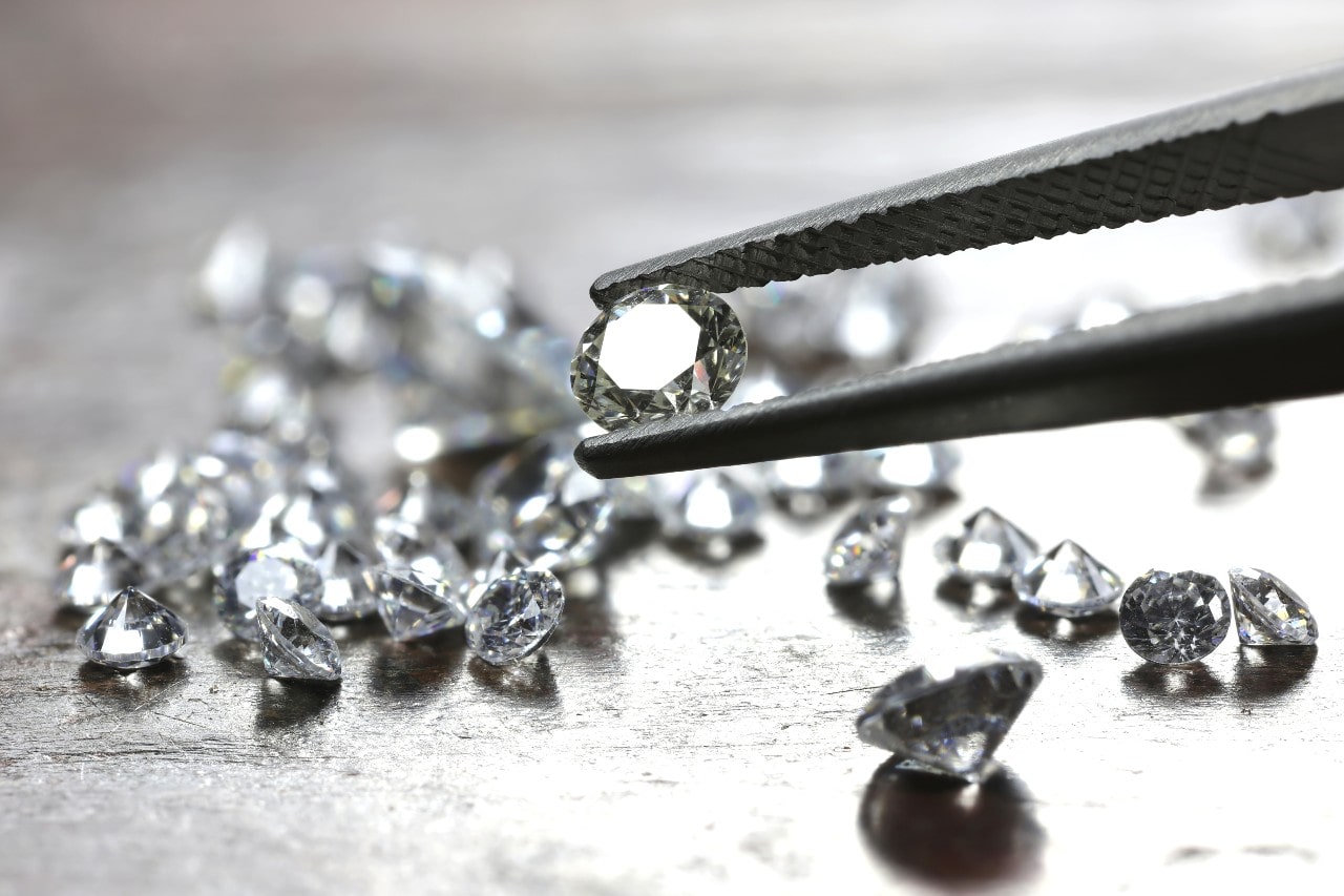 A close up of plenty of loose diamonds scattered on a wooden surface, while tweezers picked one of them. A close up of plenty of loose diamonds scattered on a wooden surface, while tweezers picked one of them.