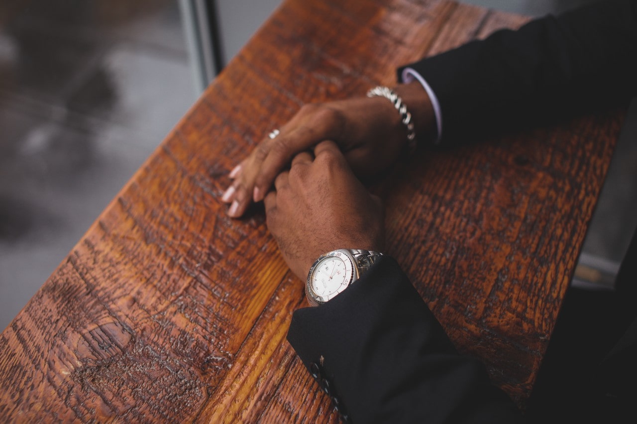 A close-up of a well-dressed man’s hands atop a wooden counter, adorned with a luxury watch, a chain bracelet, and a matching ring. A close-up of a well-dressed man’s hands atop a wooden counter, adorned with a luxury watch, a chain bracelet, and a matching ring.