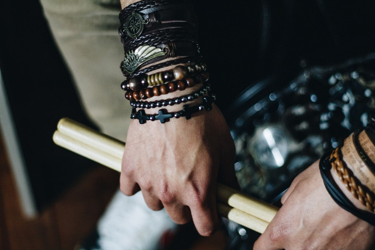 A close-up of a man’s hands holding a pair of drumsticks, adorned in multiple bracelets. A close-up of a man’s hands holding a pair of drumsticks, adorned in multiple bracelets.