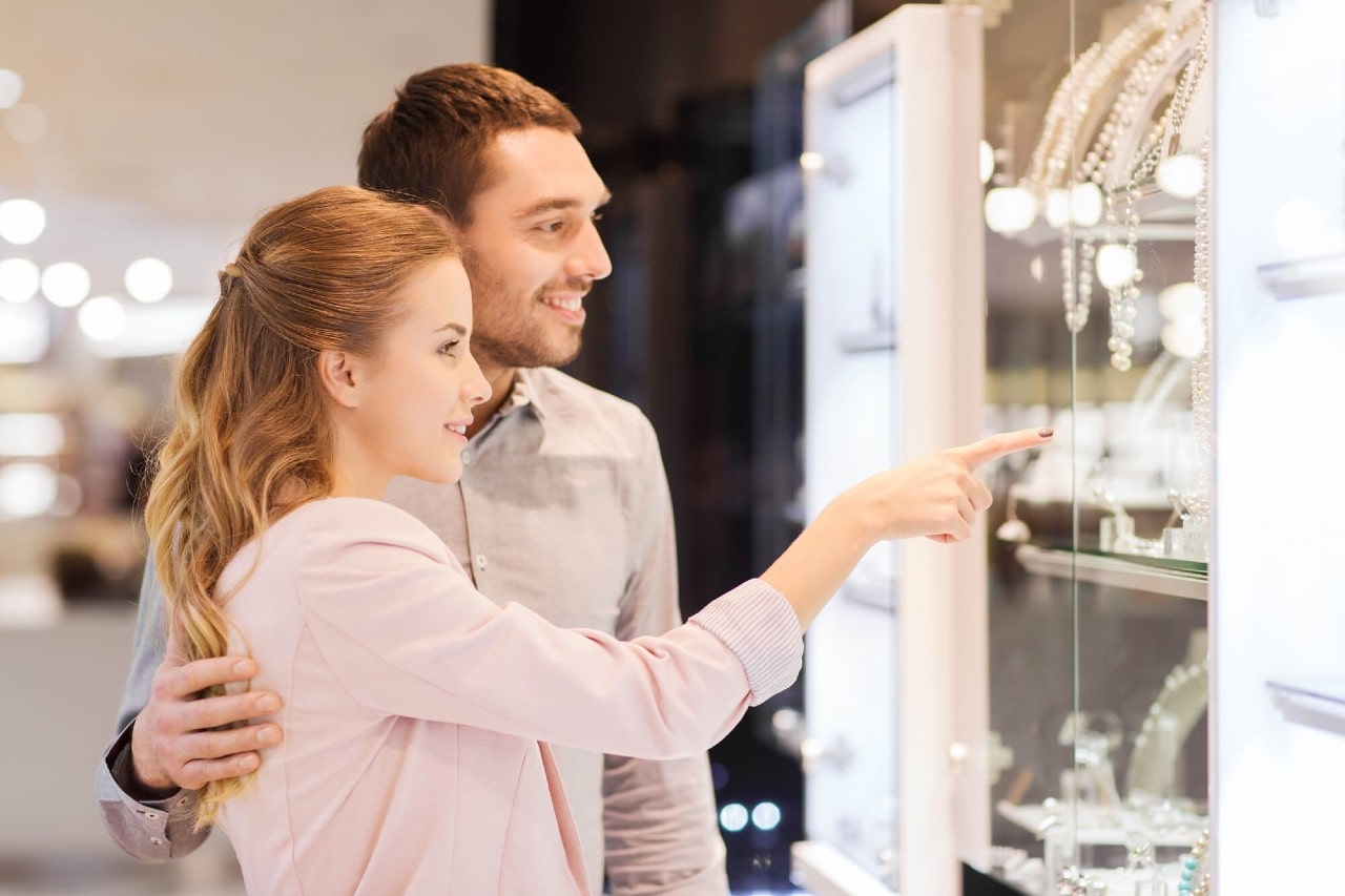 Smiling couple browsing store window; woman points at a display while her partner stands behind her, hand gently resting on her back. Smiling couple browsing store window; woman points at a display while her partner stands behind her, hand gently resting on her back.